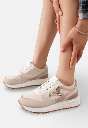 Baskets de sport beiges présentant un mélange de matériaux lisses et texturés, des semelles en caoutchouc, des lacets et des accents métalliques sur les côtés.