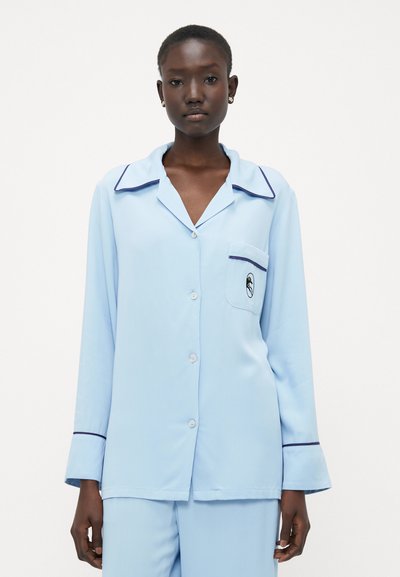 Maglia da pigiama azzurro chiaro con maniche lunghe, colletto ampio e tasca a toppa. Presenta piping blu scuro lungo i bordi del colletto e della tasca.