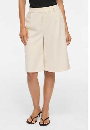 Femme portant un short beige sur mesure longueur genou, avec un top noir et des sandales à talons noires sur un fond uni.