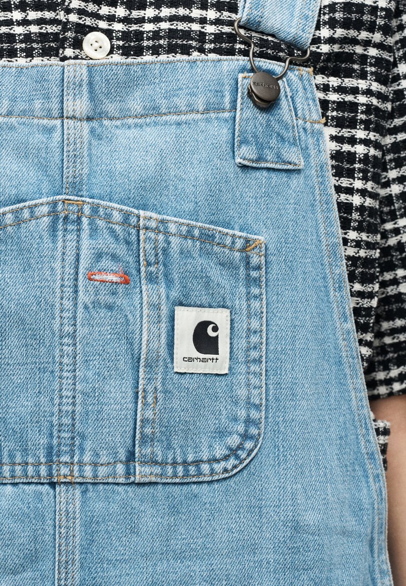 Salopette en denim bleu clair avec une poche avant affichant une étiquette de la marque Carhartt, portée sur une chemise à carreaux noir et blanc.