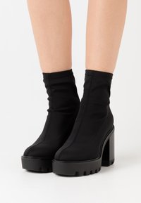 Botines negros de tobillo hechos de tela elástica, con un tacón grueso y suela con plataforma, con costuras visibles y un acabado suave.