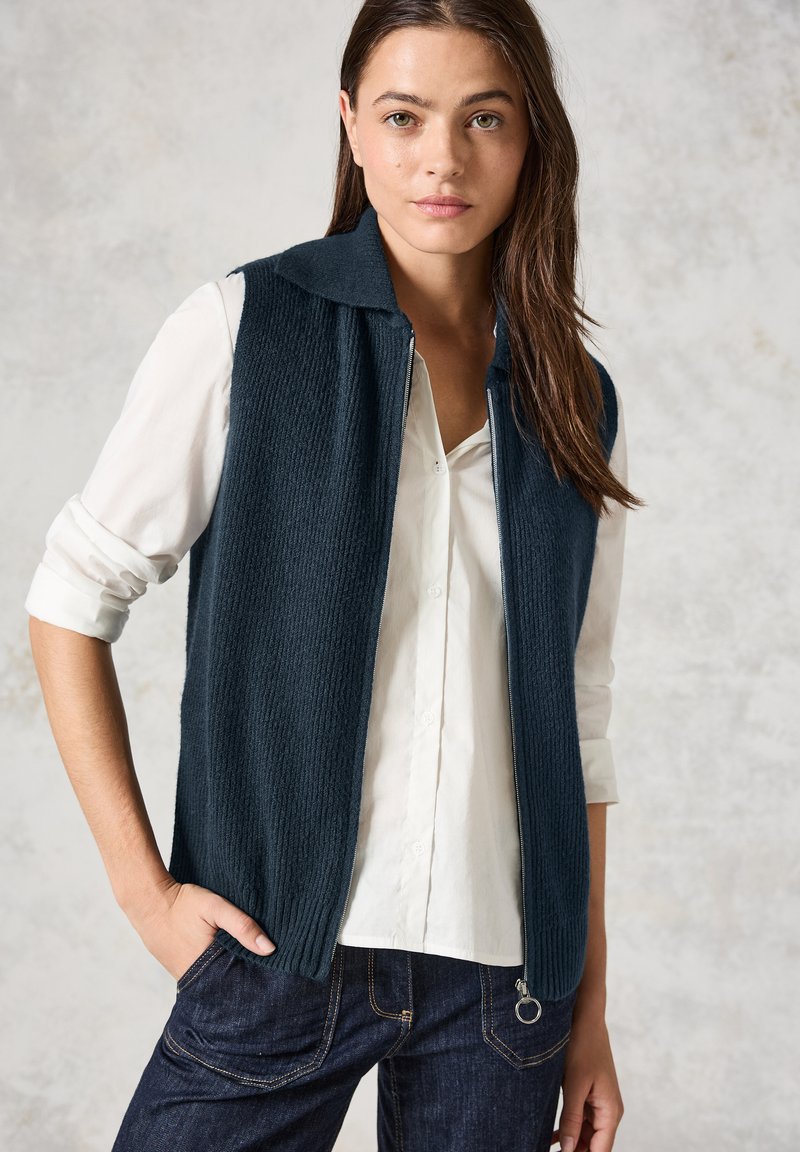 Gilet côtelé bleu marine avec un col et une fermeture éclair sur le devant, superposé sur une chemise blanche. Le gilet présente un tissu texturé et des poches latérales.