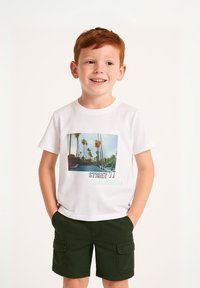 T-shirt bianca con grafica di una palma e testo "STREET 11 CALIFORNIA", abbinata a pantaloni cargo verdi. Bambino che mostra un'espressione neutra.