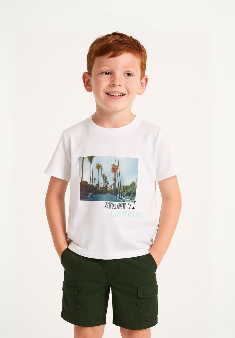 T-shirt bianca con grafica di una palma e testo "STREET 11 CALIFORNIA", abbinata a pantaloni cargo verdi. Bambino che mostra un'espressione neutra.