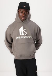 Gray hooded sweatshirt with a large white logo on the front and a front kangaroo pocket. Paired with a black cap and dark pants.