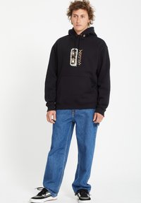 Zwarte hoodie met een voorzak, grafisch ontwerp op de borst. Gecombineerd met losse blauwe denim jeans en zwarte sneakers.