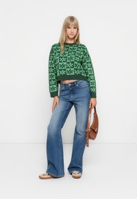 Green cropped sweater with floral patterns, paired with light blue flared jeans. Accessories include a brown shoulder bag and beige shoes.