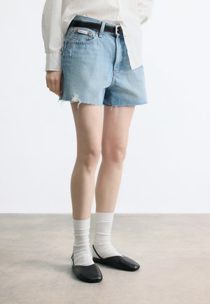 Person trägt hellblaue, abgenutzte Jeans-Shorts mit schwarzem Gürtel, weißes Langarmshirt, weiße Socken und schwarze flache Schuhe.