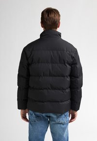Uomo con giacca imbottita nera e jeans blu, in piedi di spalle su uno sfondo bianco uniforme.