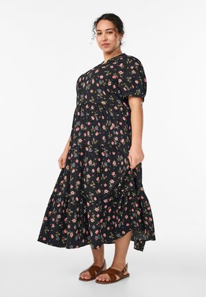 Naine kannab musta lillimustriga midi-kleit, puffvarrukatega ja pruune avatud varvastega sandaale, seisab ühevärvilise valge tausta ees.