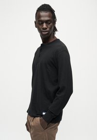 Chemise henley noire à manches longues en tissu texturé, avec trois boutons au col et une petite étiquette logo à la taille.