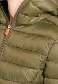 Olijfgroene pufferjas met horizontale stiksels, een capuchon en een zilveren ritssluiting met een oranje trekker. Soepele, gewatteerde structuur.