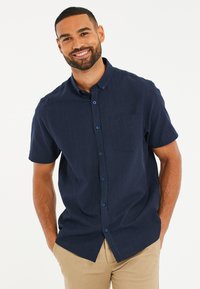 Camisa de manga corta, de color azul marino, con botones, hecha de tela texturizada, que incluye un bolsillo en el pecho y botones azules en contraste.