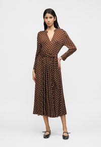 Hnedé zavinovacie šaty s bodkami, dlhé rukávy, výstrih do V a pás s opaskom. Prúdiaca silueta so dĺžkou do polovice lýtok. Jednoduché čierne balerínky.