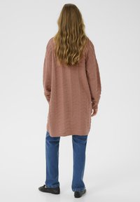 Femme aux cheveux longs et ondulés portant un cardigan oversize texturé rose poussiéreux, un jean bleu et des chaussures noires, se tenant de dos sur un fond blanc.
