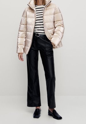 Kobieta w błyszczącej beżowej pikowanej kurtce, czarnych skórzanych spodniach z paskiem, czarnych mokasynach oraz czarno-białej paskowanej koszulce z długim rękawem.