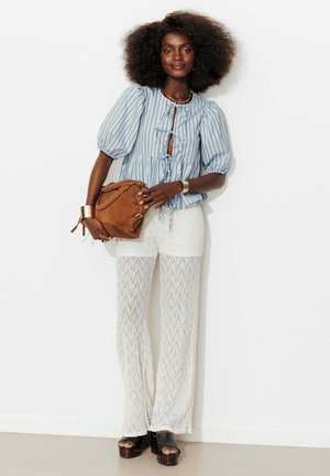 Femme avec coiffure afro portant un chemisier rayé bleu, un pantalon en dentelle blanc, des sandales à plateforme noires, et tenant un sac en daim marron.