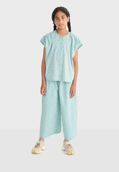 Fille aux cheveux tressés portant un haut sans manches bleu clair à motifs et un pantalon ample, debout avec une expression neutre sur un fond clair.