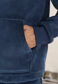Felpa in misto cotone blu navy con texture a coste, dotata di tasca frontale e polsini a coste. Vista ravvicinata di una mano nella tasca.