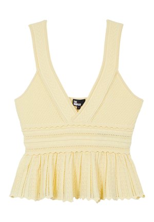 Femme portant des lunettes de soleil, un haut en maille jaune sans manches, un jean bleu et des sandales, tenant une veste en cuir marron dans une main.