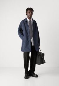 Mornarska vodootporna jakna s patentnim zatvaračem, uparena s bijelom košuljom i sivom kravatom. Crne hlače i elegantne cipele upotpunjuju izgled.