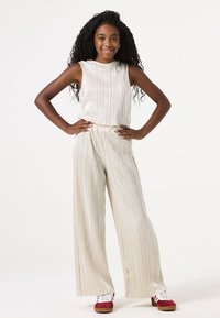Lichtbeige twee-delig outfit met verticale plooien; mouwloze top en wijde broek. Draag met rode schoenen en witte veters.