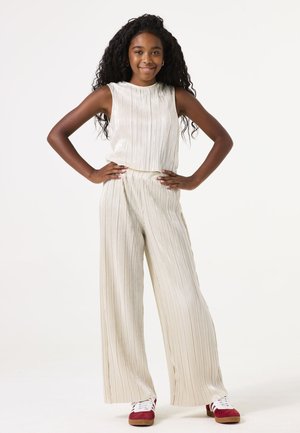 Conjunto de dos piezas en beige claro con plisados verticales; blusa sin mangas y pantalones de pierna ancha. Llevado con zapatos rojos y cordones blancos.