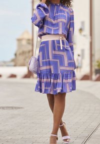 Žena v fialových šatech s geometrickým vzorem, se skládaným lemem, béžovým opaskem v pase, levandulovou kabelkou a ladícími sandály na podpatku, jde po dlažební ulici.