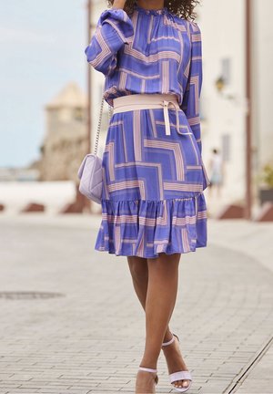 Donna in un abito viola con motivo geometrico e orlo arricciato, cintura beige in vita, borsa lavanda e sandali con tacco coordinati che cammina su una strada pavimentata.