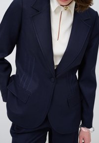 Blazer blu navy con revers classico, due tasche e dettagli decorativi in cucitura. Abbinato a una camicia bianca e a un braccialetto trasparente.