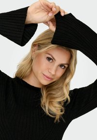 Blonde Frau mit lockeren Locken trägt einen schwarzen gerippten Pullover mit weiten Glockenärmeln und hebt die Hände über ihren Kopf vor einem schlichten Hintergrund.