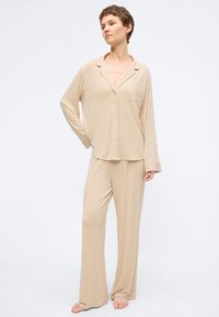 Γυναίκα όρθια, ξυπόλυτη, φορώντας χαλαρό, μπεζ πουκάμισο με μακριά μανίκια και κουμπιά και ταιριαστό φαρδύ παντελόνι, μπροστά σε απλό λευκό φόντο.