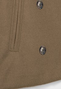 Gros plan d'un manteau en tissu marron avec deux boutons foncés et une poche latérale verticale avec détail de couture.