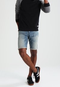 Camisa de manga larga negra y gris combinada con pantalones cortos de mezclilla azul claros, mostrando un ajuste relajado y puños enrollados. Zapatos negros con rayas blancas.