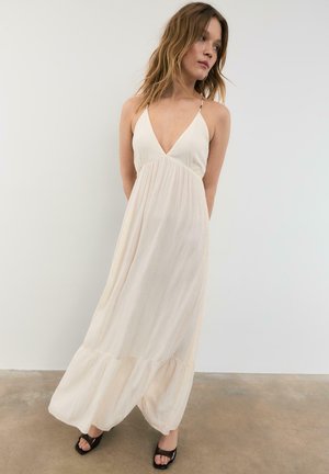 Młoda kobieta w kremowej, bezrękawnikowej sukience maxi z głębokim dekoltem w serek, stojąca boso na betonowej podłodze na tle gładkiej białej ściany.