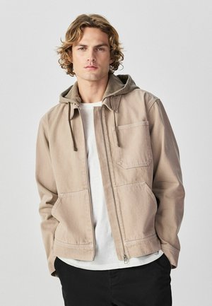 Young man with wavy blonde hair wearing a beige zip-up jacket with a hood over a white T-shirt and black pants.
