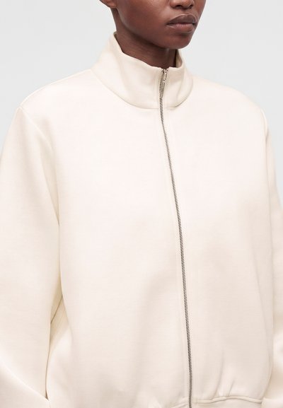 Personne portant une veste crème zippée avec un col montant sur un fond clair uni.
