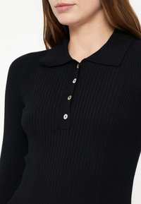 Femme portant un haut noir côtelé à manches longues avec un petit col évasé et quatre boutons nacrés visibles sur la poitrine.