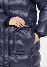 Marinblå puffjacka med en kviltad design och en synlig ficka, blank yta och svart innerfoder. Knäppning vid fickan.