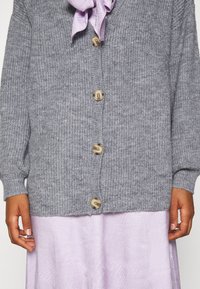 Gray knitted cardigan with seven buttons and ribbed texture. Features a collared neckline with a light purple scarf. Worn over a pale purple dress.