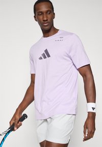 Jasnofioletowa koszula z krótkim rękawem z ciemnoszarym logo Adidas i napisem z przodu, wykonana z miękkiego materiału, noszona z białymi szortami.