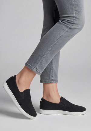 Persoon draagt zwarte slip-on gebreide sneakers met witte zolen en grijze nauwsluitende spijkerbroek tegen een effen lichte achtergrond.