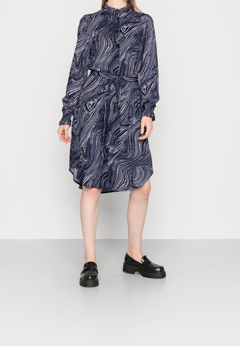 Tummansininen kuvioitu mekko, jossa on pitkät hihat, korkea kaulus ja vyötärösolmu; sisältää virtaavaa kangasta, jossa on aaltoileva valkoinen kuvio ja polvenpituinen helma.