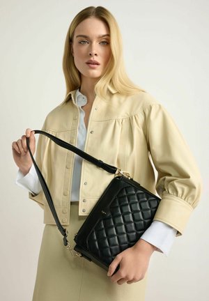 Blondynka w beżowej kurtce i spódnicy trzyma czarną pikowaną torebkę za pasek na ramię na jednolitym jasnym tle.