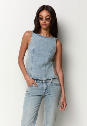 Frau trägt hellblaues ärmelloses Denim-Top und Jeans, mit Sonnenbrille, steht vor einem schlichten weißen Hintergrund, eine Hand in der Tasche.