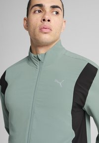 Lichtgroene zipjack met zwarte accenten en een Puma-logo op de borst. De stof heeft een gladde textuur en een gestructureerde pasvorm.