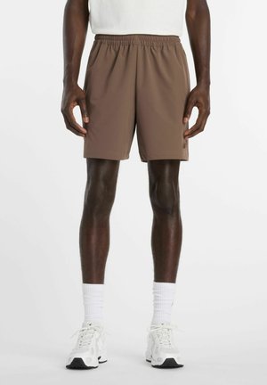 Homme portant un short marron à taille élastique, des chaussettes blanches montantes et des chaussures de sport blanches, debout sur un fond blanc.