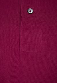 Camisa polo color burdeos con una textura suave, que presenta un botón plateado y un sutil detalle acanalado cerca del cuello y en la zona del bolsillo.