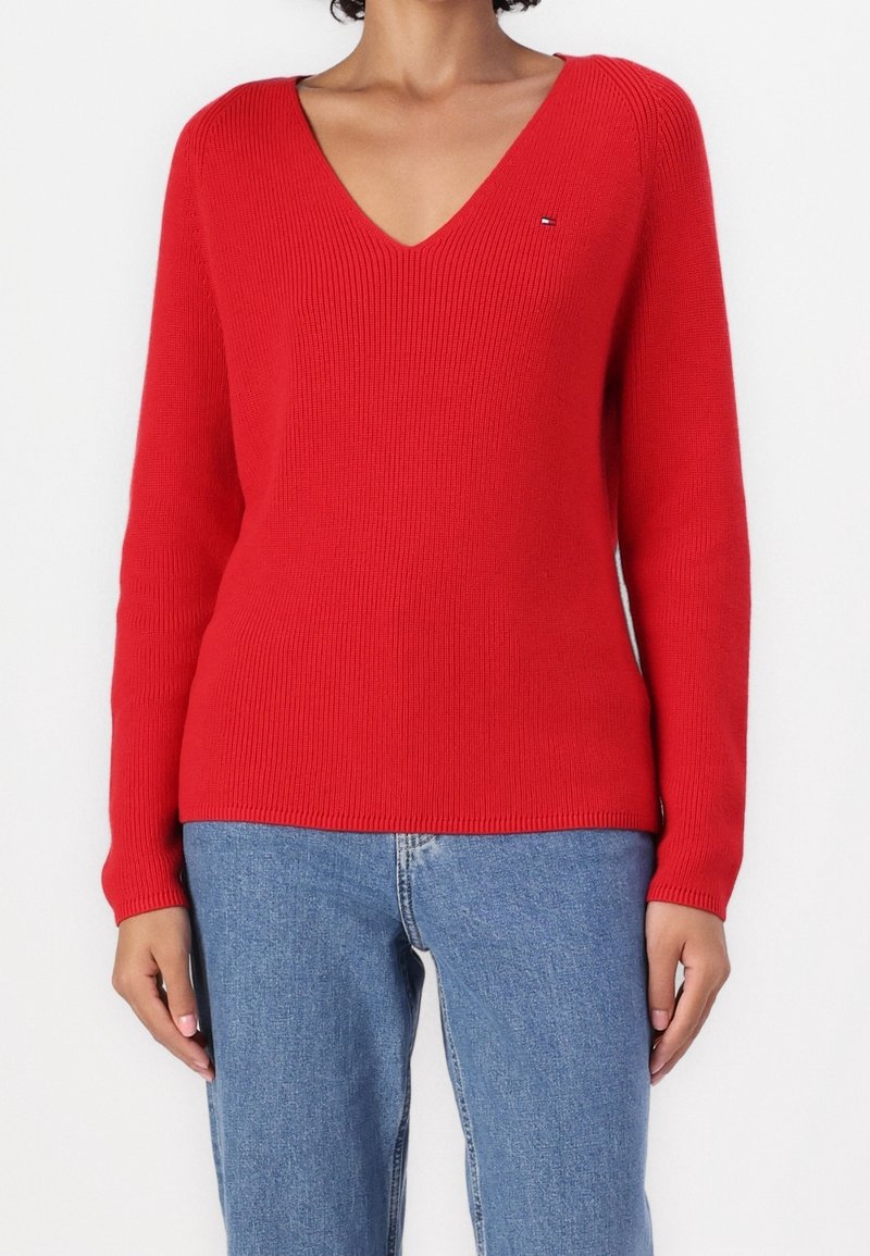 Maglione rosso lavorato a maglia con scollo a V, maniche lunghe, trama a coste e piccolo logo sul petto. Abbinato a jeans di jeans blu.