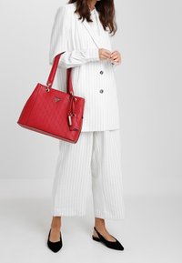 Rode gestructureerde handtas met geometrisch patroon en zilverkleurige hardware, vastgehouden door een persoon in een wit gestreept pak en zwarte schoenen.